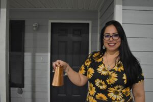 Selma Belcher holding a cowbell outside of Habitat’s model home. The team rings the bell each time an applicant qualifies for the Habitat program.